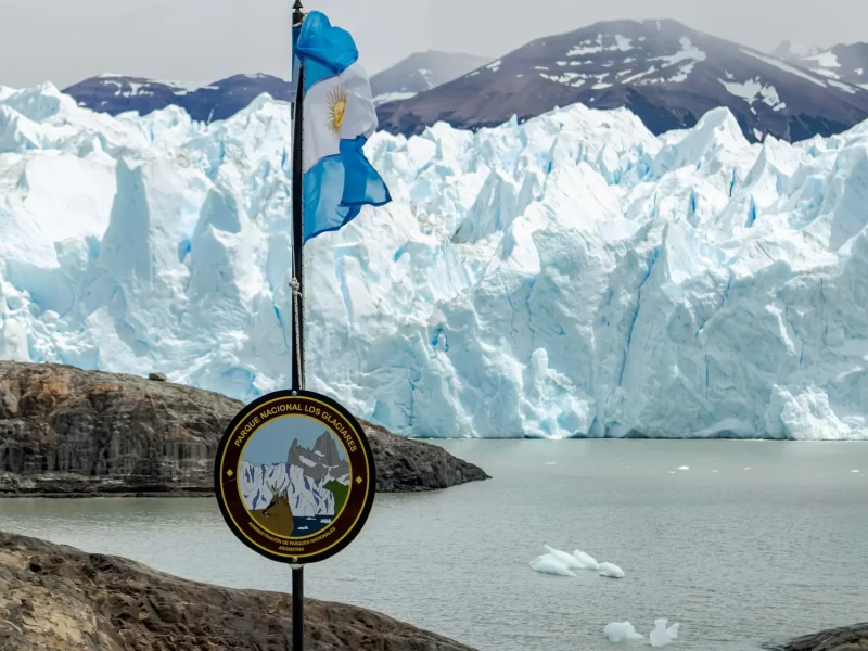 Los Glaciares National Park sign with the Argentinian flag in the foreground and the massive ice walls of the Perito Moreno Glacier in the background.