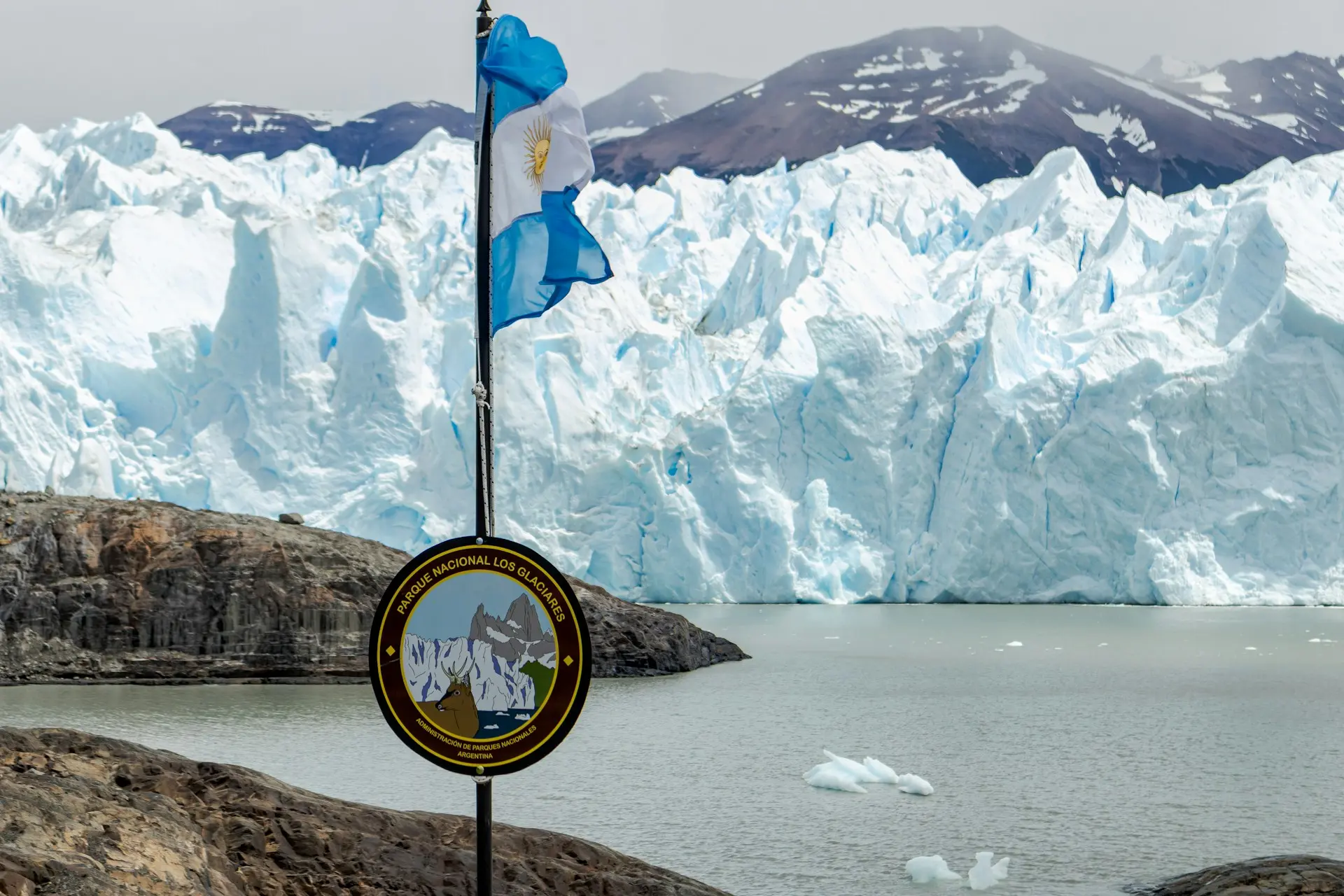 Los Glaciares National Park sign with the Argentinian flag in the foreground and the massive ice walls of the Perito Moreno Glacier in the background.