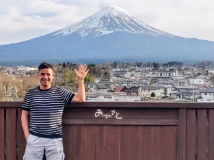 Man waving in front of the snow-capped Mount Fuji and town view from a wooden viewpoint in Fujikawaguchiko, Japan.