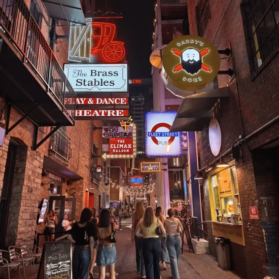 Printers Alley in Nashville illuminated at night with vibrant neon signs for The Brass Stables and Fleet Street Pub as people walk down the alley.