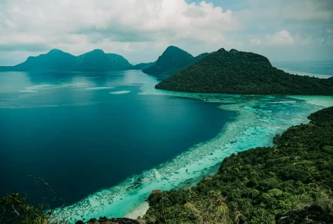 An aerial, high-angle photograph of Tun Sakaran Marine Park in Malaysia, showcasing mountainous, rainforested islands and the extensive shallow, clear turquoise coral reefs and deep blue waters under a cloudy sky