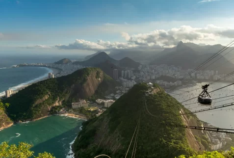 A cable car ascending Sugarloaf Mountain, offering a panoramic aerial view of Rio de Janeiro's city skyline, beaches, and lush green mountains.