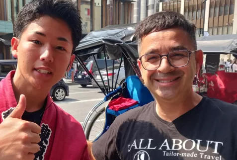 young Japanese rickshaw driver in traditional red clothing giving a thumbs up, smiling next to a Caucasian man wearing an 'All About Tailor-made Travel' t-shirt on a city street in Japan, with a rickshaw in the background